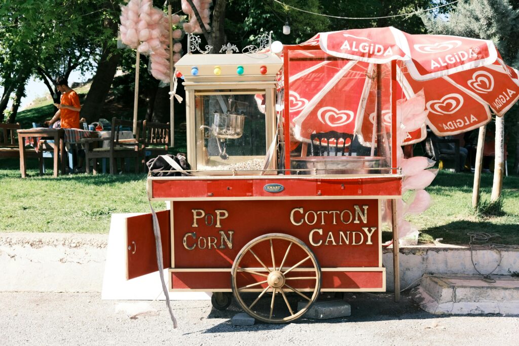 Red popcorn and cotton candy stall in a sunny park setting with people enjoying a day out.